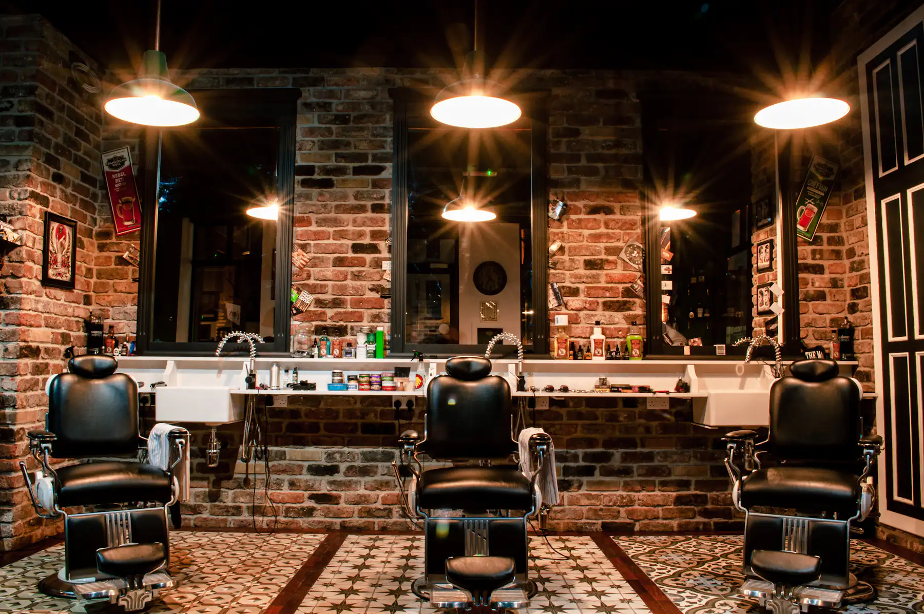 Three black leather barber chairs under pendant lights in front of a brick wall